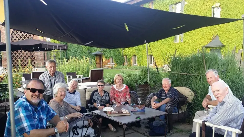 A group of smiling seniors seated outdoors under a canopy, enjoying drinks and snacks in a garden courtyard.