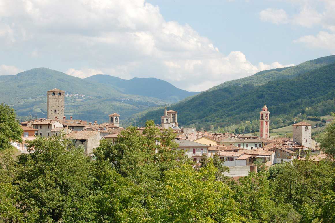 A charming hill town with terracotta roofs and stone bell towers is nestled among forested mountains.