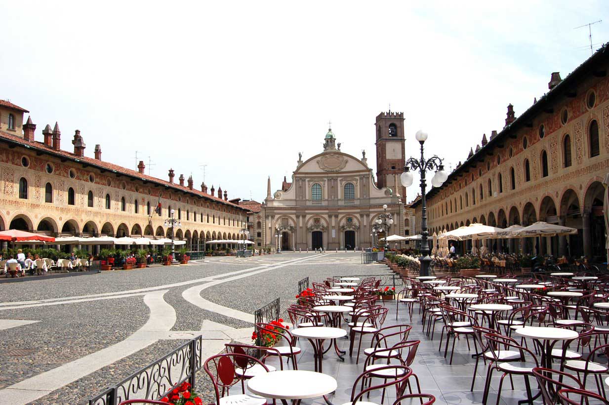 A grand Renaissance square lined with arched arcades and outdoor cafés leads to an ornate church facade with a bell tower.