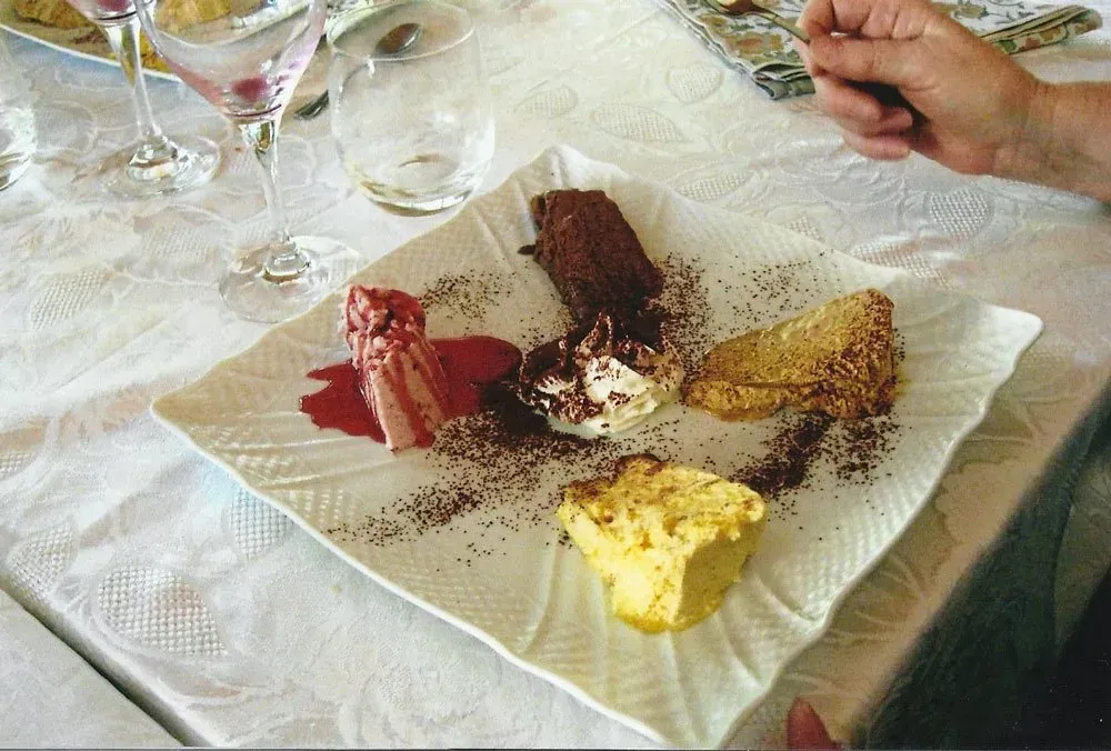 Plate of assorted desserts including chocolate cake, tiramisu, semifreddo, and fruit sauce on a white tablecloth.