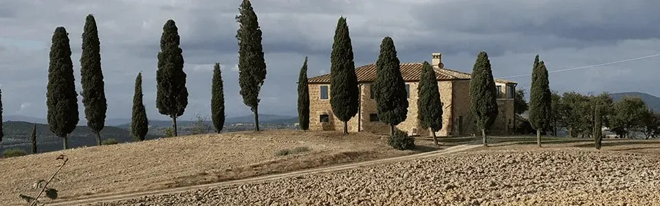 A rustic stone farmhouse surrounded by tall cypress trees on a dry, rolling landscape under a cloudy sky.