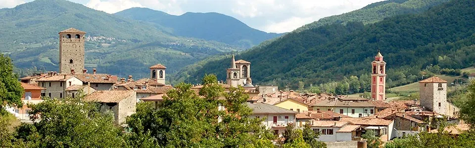 A charming hillside village with historic stone towers, red-tiled roofs, and lush green mountains in the background.