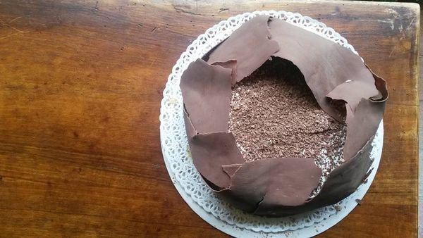 A chocolate cake topped with dark chocolate shards and grated chocolate, set on a lace doily over a wooden surface.