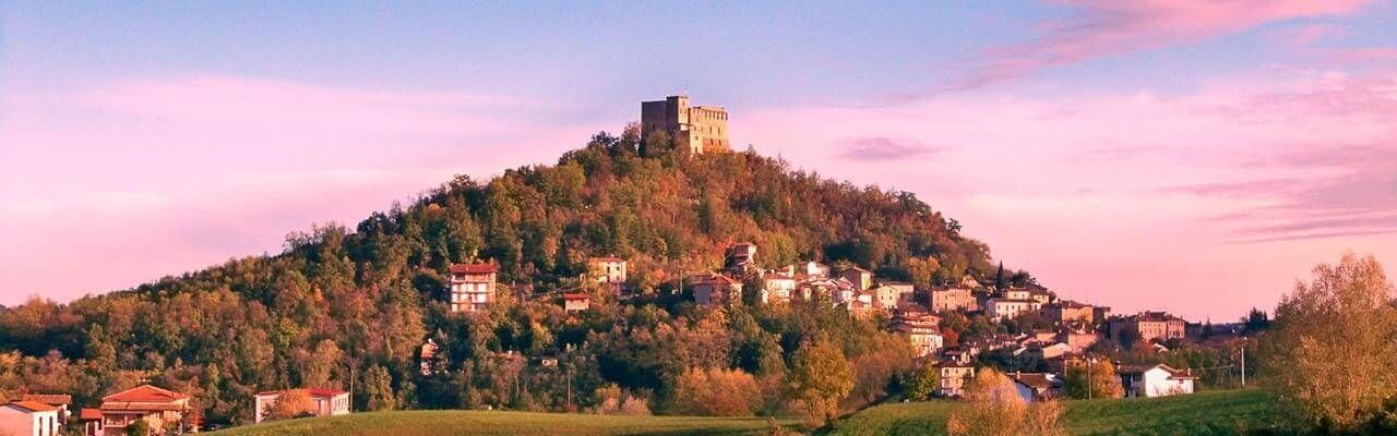 Panoramic view of a hilltop village with a prominent castle at sunset, under a pink sky.