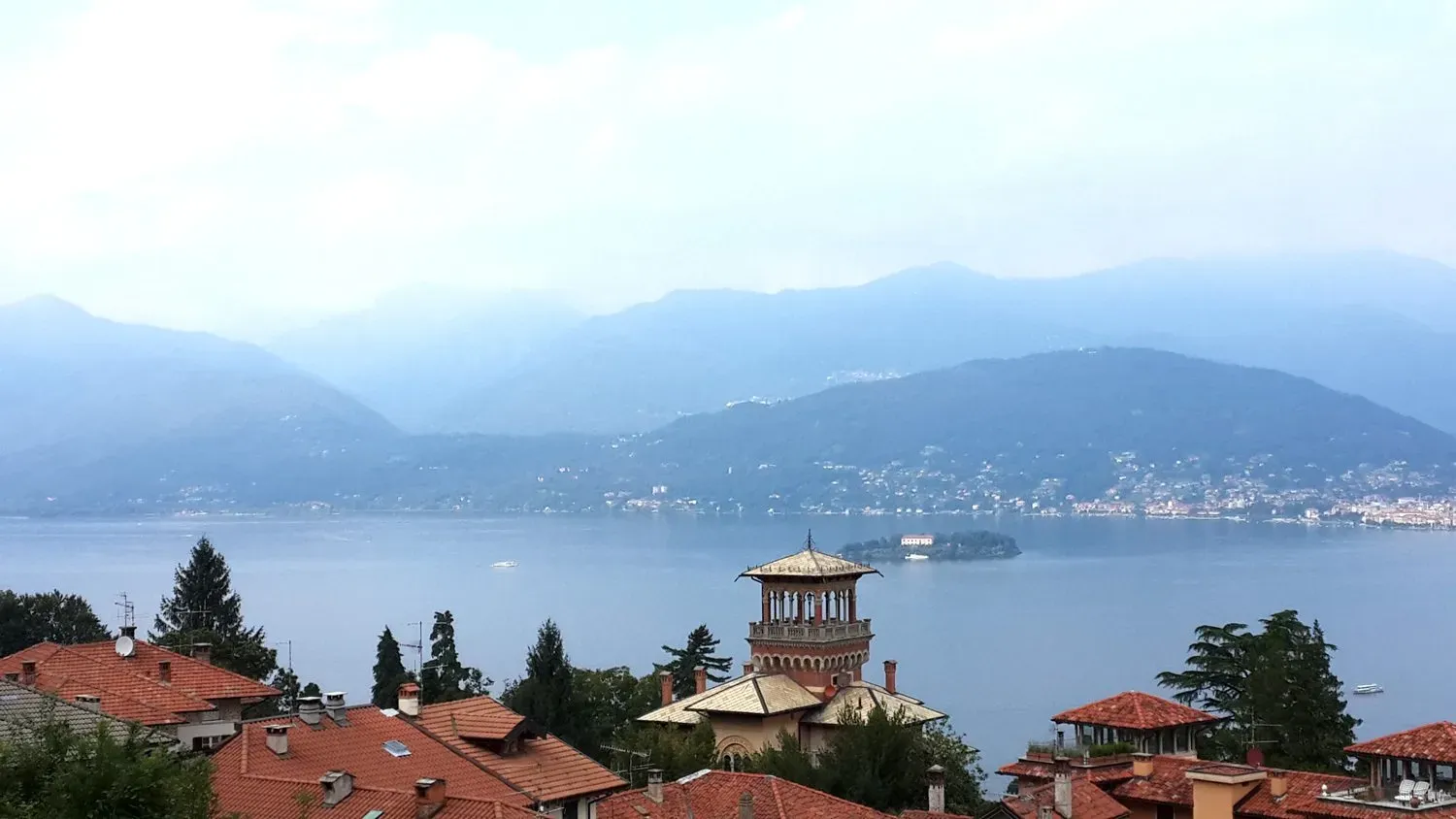 Lake Maggiore view—terracotta rooftops, a grand turreted villa, hazy blue mountains, and calm water dotted with boats.