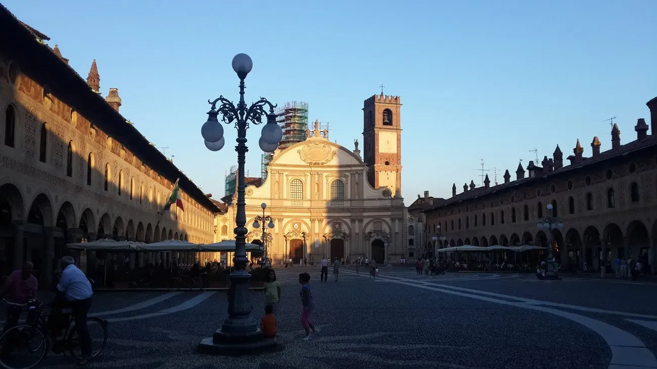 Sunlit piazza in Vigevano, Italy, with a cathedral and tower framed by arcaded buildings.