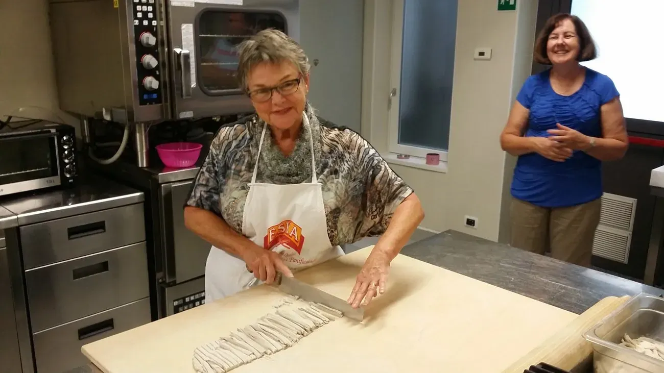 A woman is cutting fresh pasta dough in a kitchen. Another woman smiles in the background.