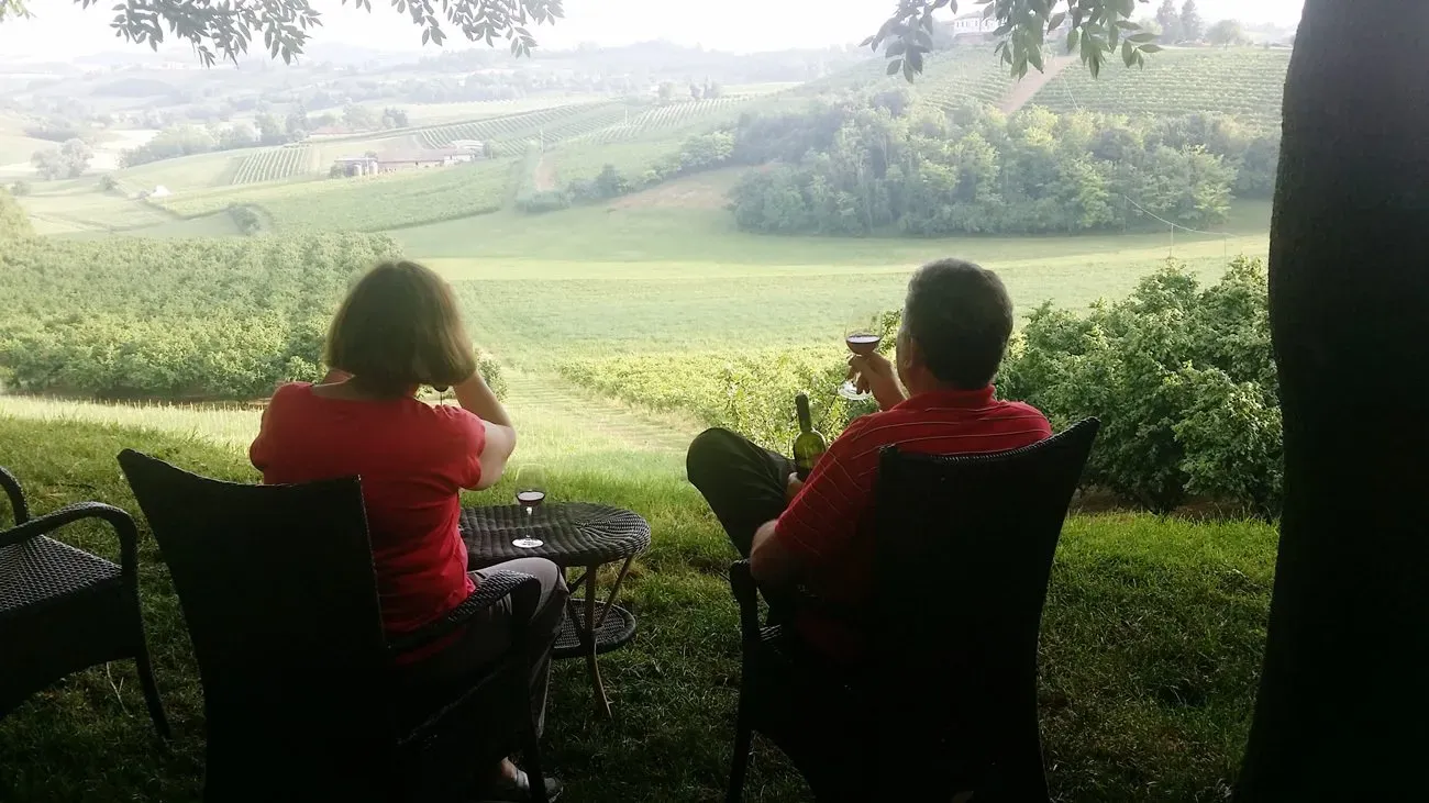 Two people in red shirts sit under tree shade, sipping wine while looking out over lush vineyard hills of Montallegro.