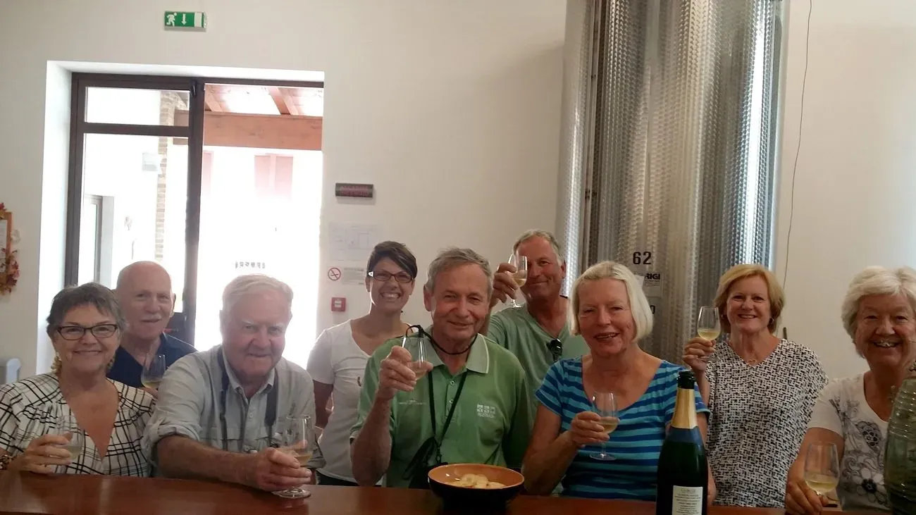 Group raising wine glasses around a table inside a winery with a stainless steel fermentation tank in the background.