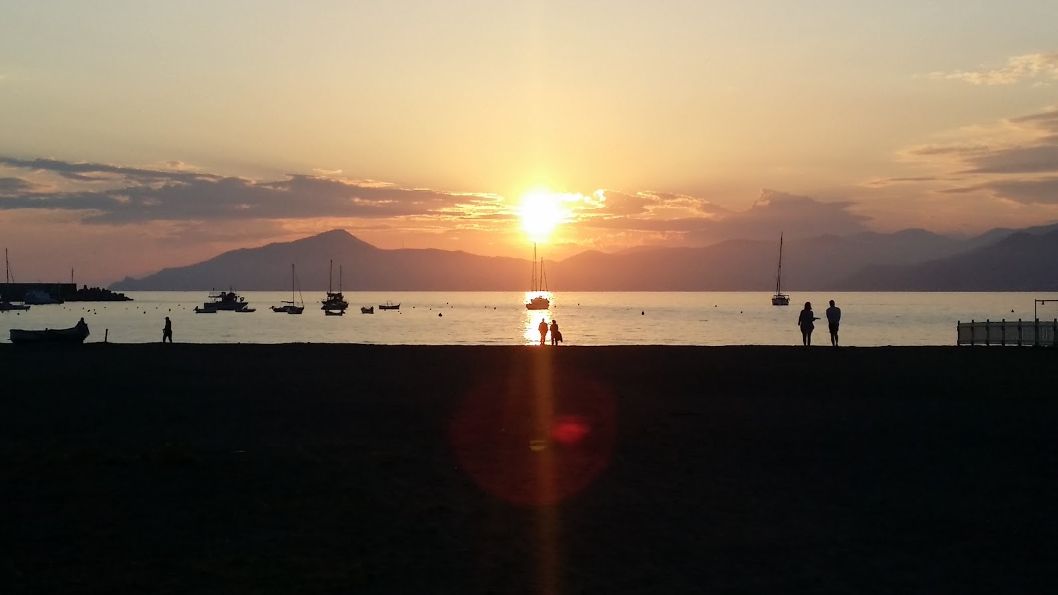 A couple walking on a beach at sunset with boats in the background.