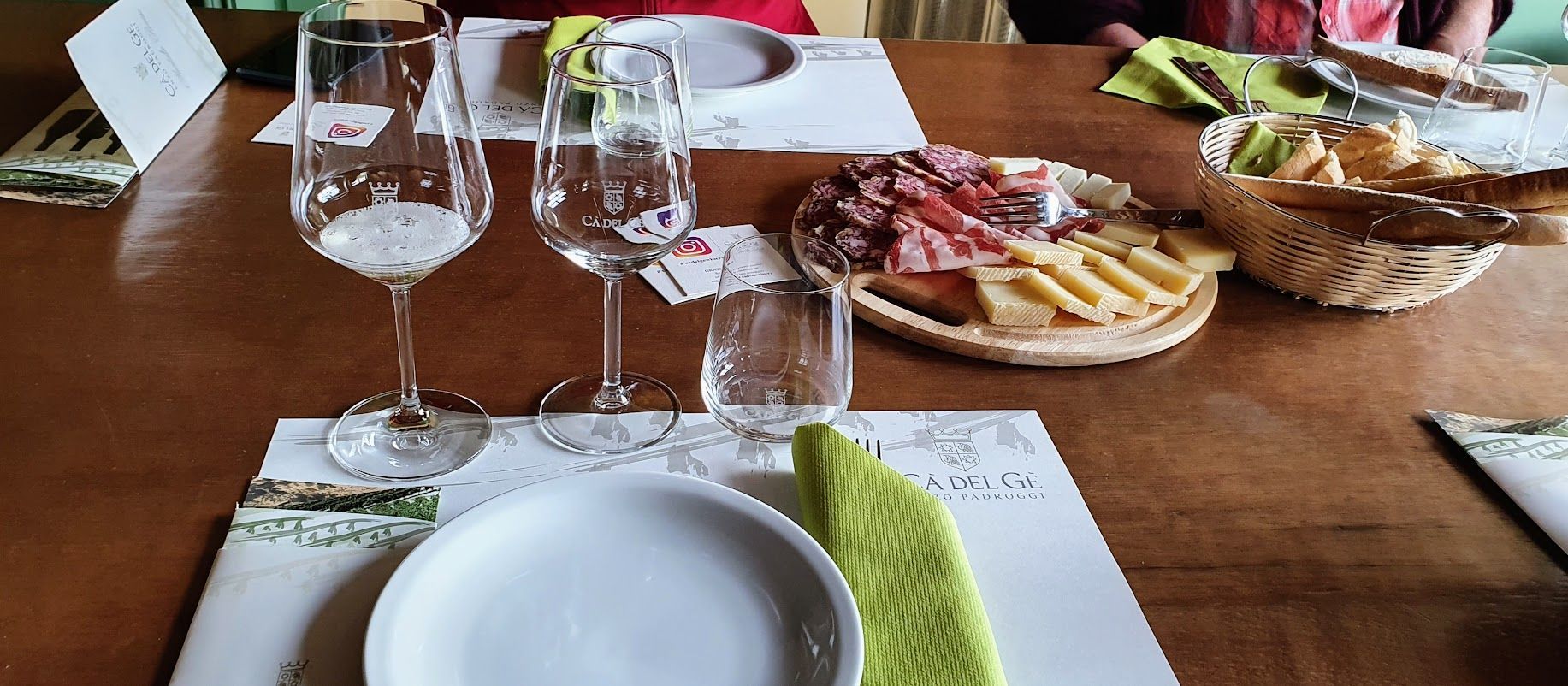 Dining table setup with empty wine glasses, a wooden platter of assorted meats and cheeses, and a basket of bread.