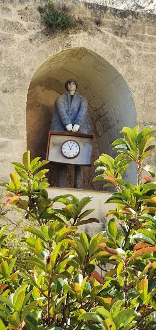 A statue of a man standing in front of a clock surrounded by plants.