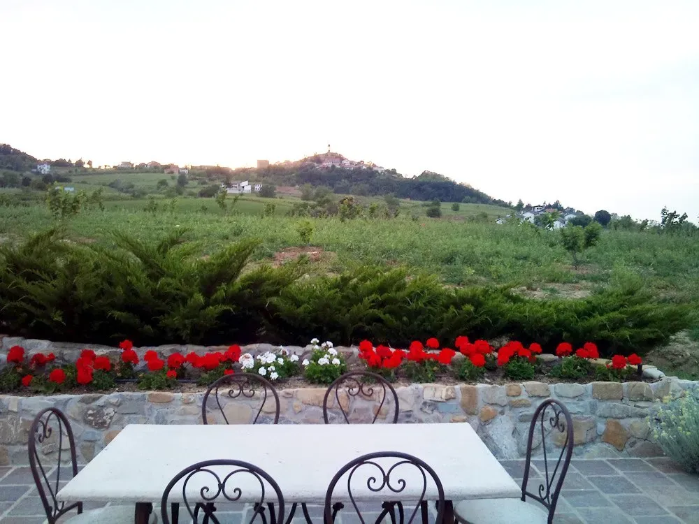 Patio with wrought iron chairs and stone table facing a flower bed and green fields with a hilltop village in the distance.