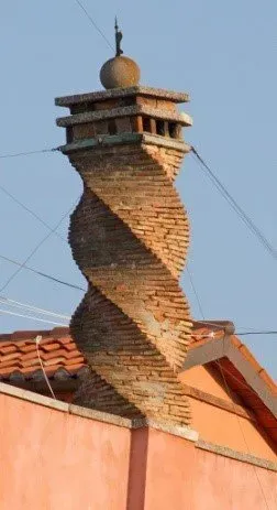 Twisting brick chimney with a spherical finial atop a terracotta rooftop.