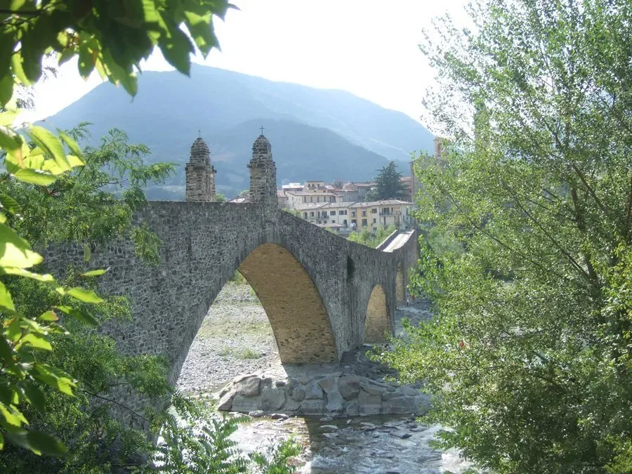 Ponte Vecchio (Devil's Bridge) in Bobbio, Italy, with a scenic village and mountains in the background.