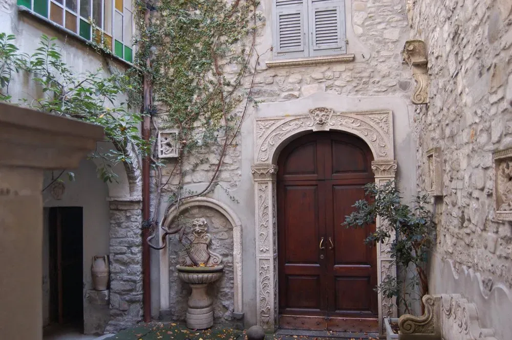Stone courtyard in Bobbio with an ornate wooden door, ivy-covered walls, and a decorative fountain.