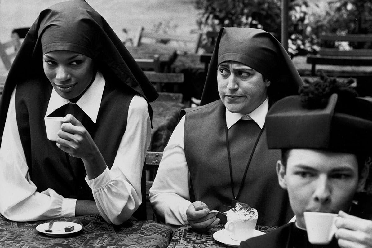 Black and white photo of two nuns and a priest in traditional attire, enjoying coffee at an outdoor café.