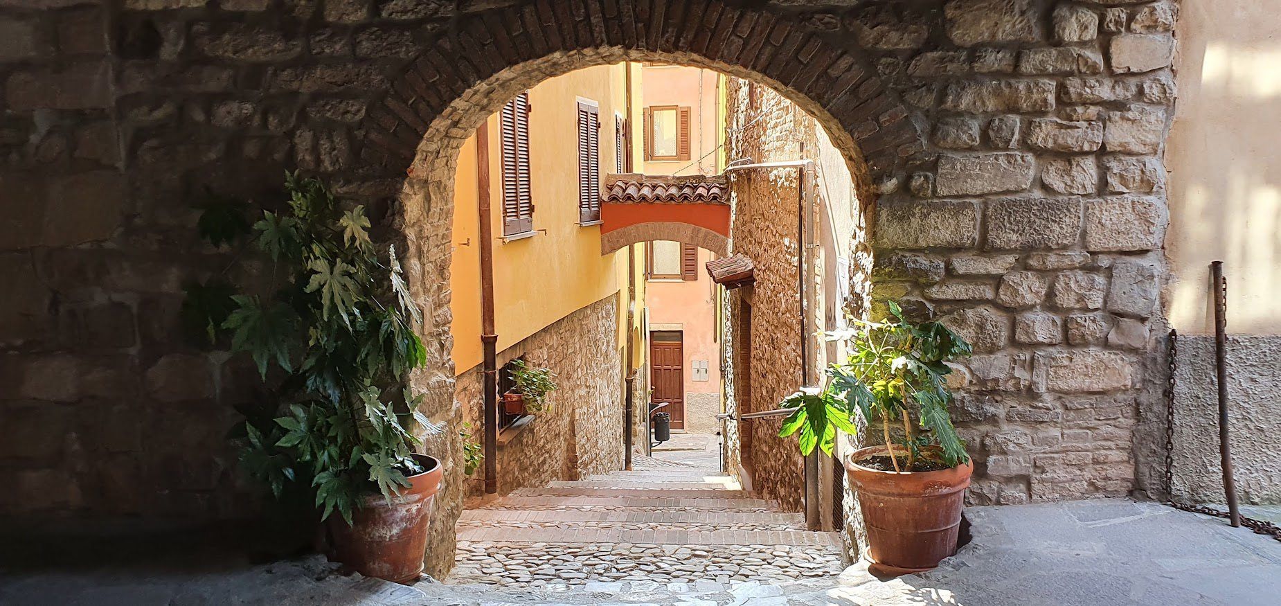 Stone archway framing a narrow, sunlit alley with colorful buildings and potted plants.