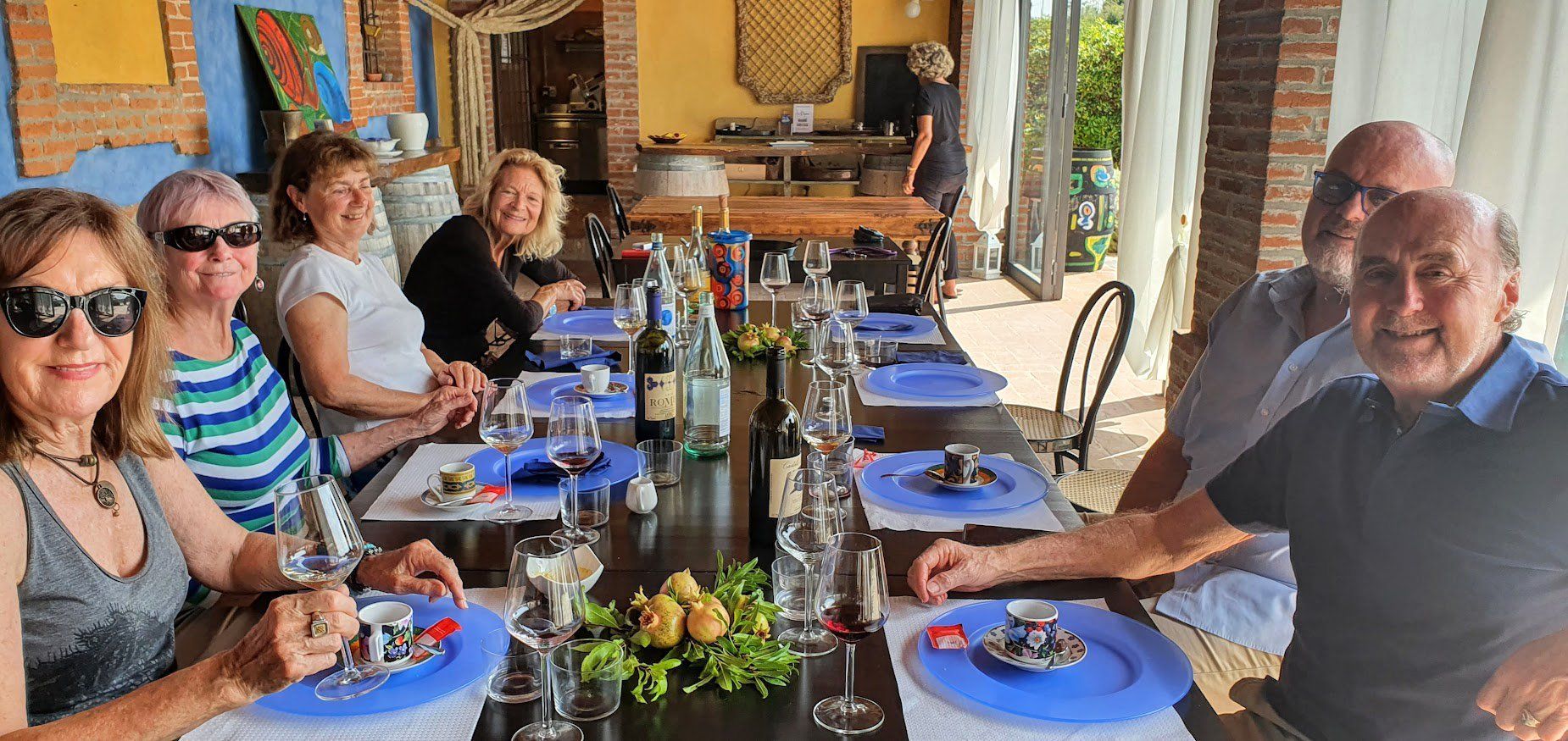 A group of smiling people seated at a long table set with blue plates, espresso cups, and wine glasses in a rustic Italian.