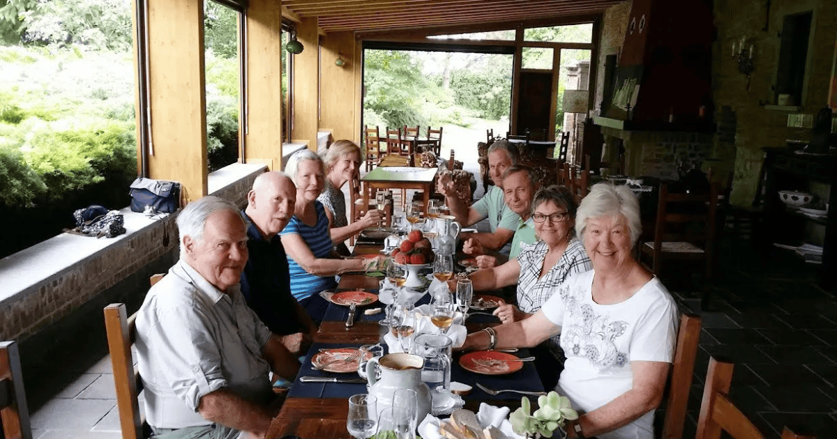 Group dining at a long table in a rustic restaurant with large windows and garden views.