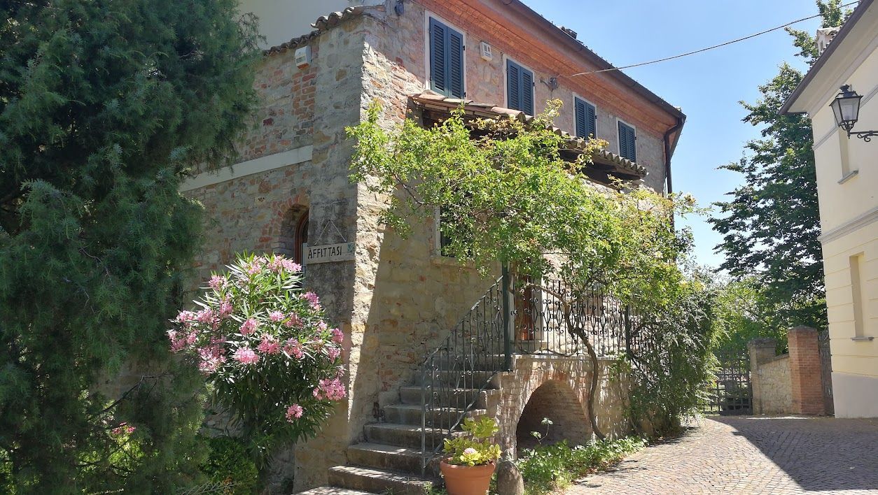 Stone house with shuttered windows, covered by greenery and pink flowers, located on a cobbled street.