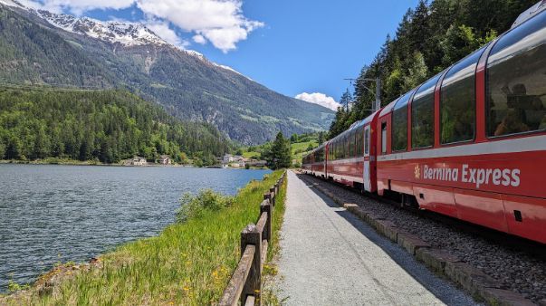 A red and white train is going down the tracks next to a lake.