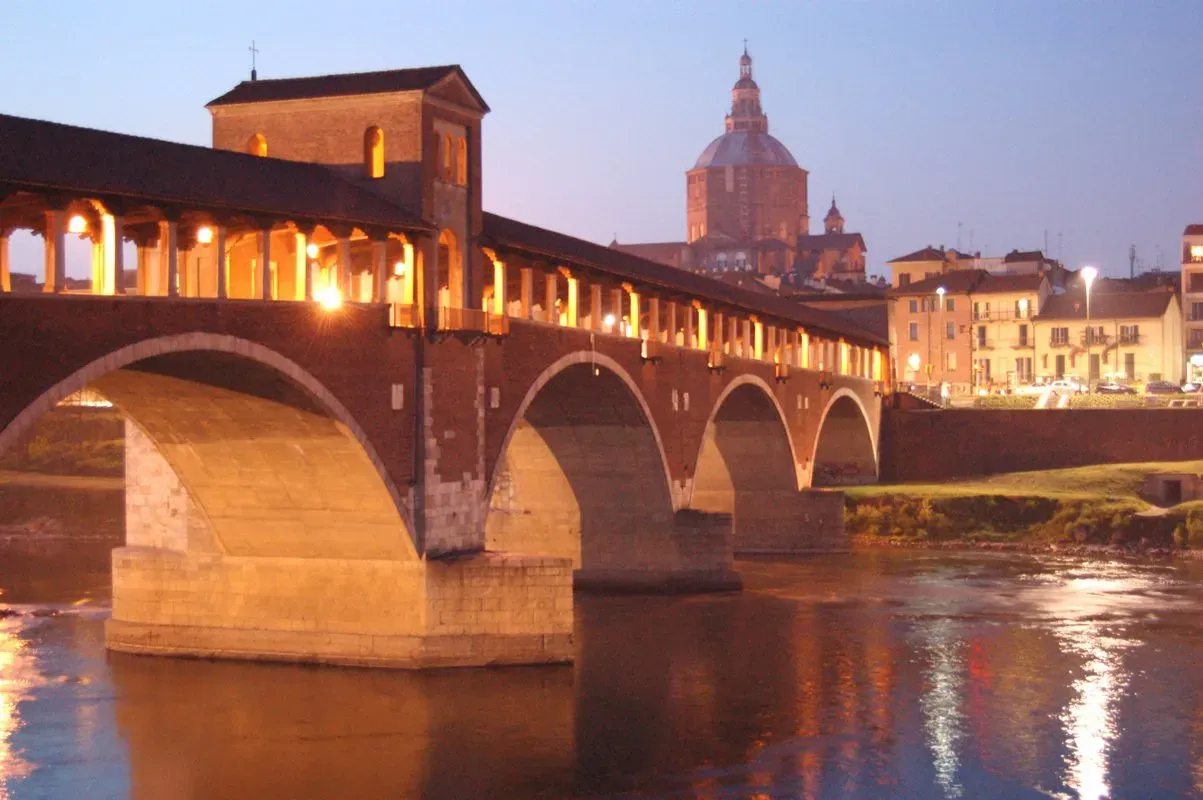 Ponte Coperto in Pavia at dusk, with lights glowing and the Duomo in the background.