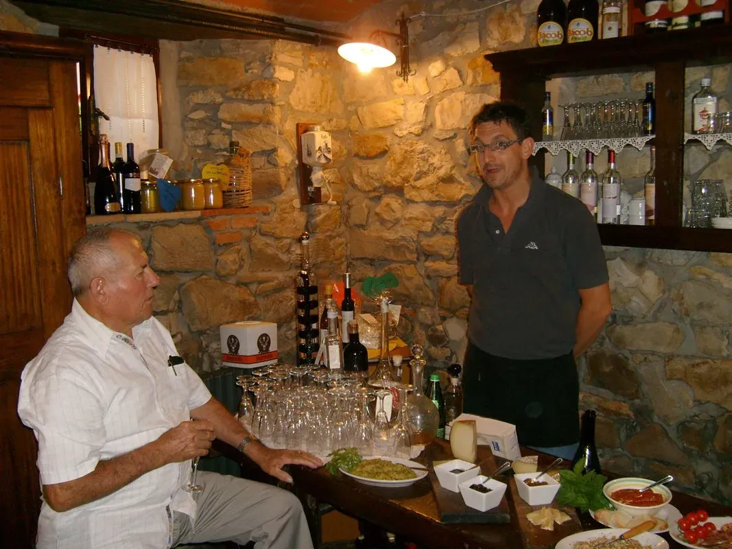 Two men talk inside a rustic bar with stone walls, surrounded by wine bottles, cheese, and glasses set for tasting.