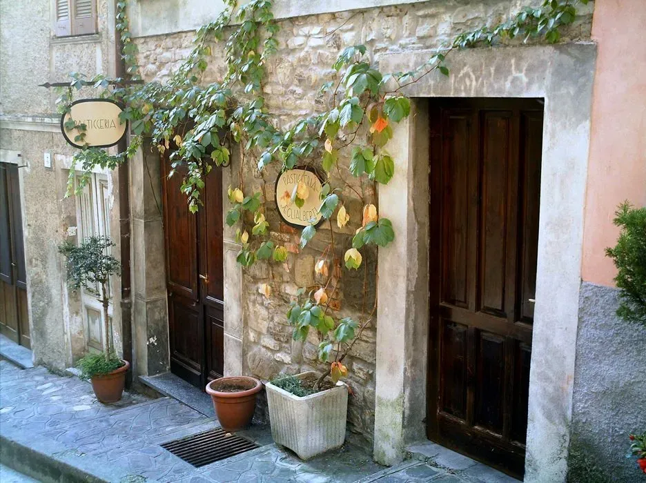 Rustic storefront in a narrow alley, with wooden doors, stone walls, potted plants, and hanging signs for a pasticceria.