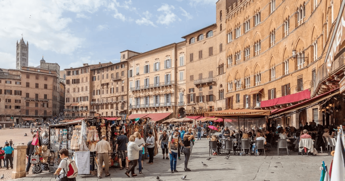 Crowds gather in Siena’s Piazza del Campo, with outdoor cafés, market stalls, and medieval buildings under a clear sky.