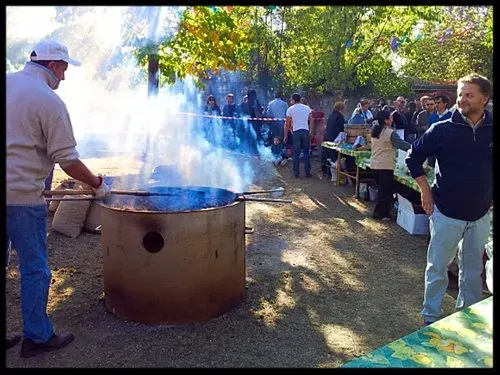 Outdoor gathering with people around a large smoking roasting pit, enjoying a rustic cooking event in a sunlit setting.