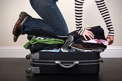 A person is struggling to close an overstuffed suitcase by pressing down on it with their hands and knees.