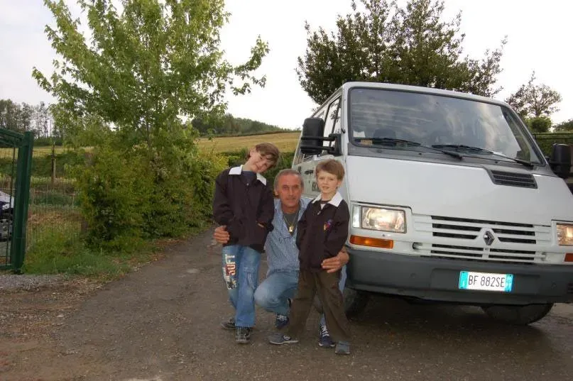 Man crouching with two boys in matching jackets beside a white Renault van on a country road.
