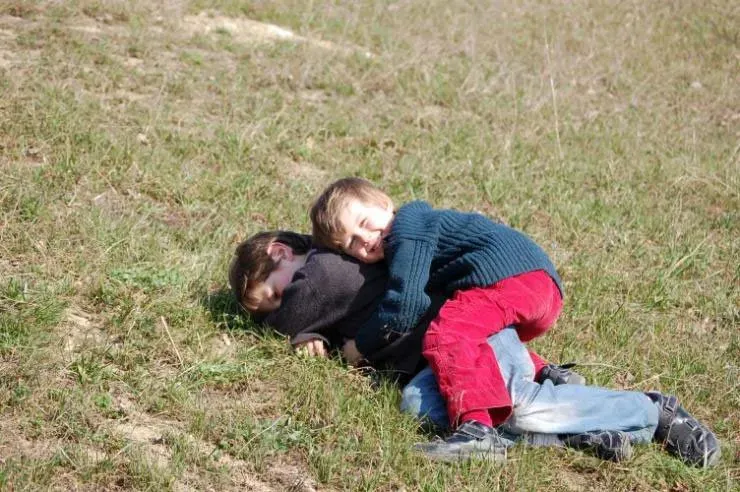 Two young boys playfully lying on a grassy hill, one hugging the other while smiling.