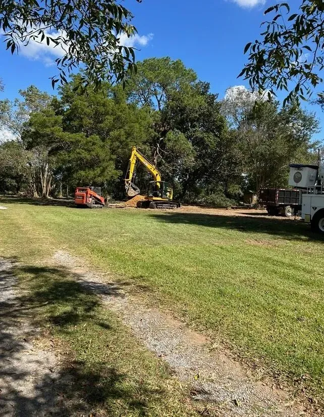 Construction site with excavator and bobcat on grassy field, trees in the background, blue sky.
