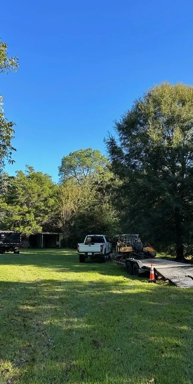 A white pickup truck and construction equipment sit on a grassy lawn with trees and a clear blue sky.