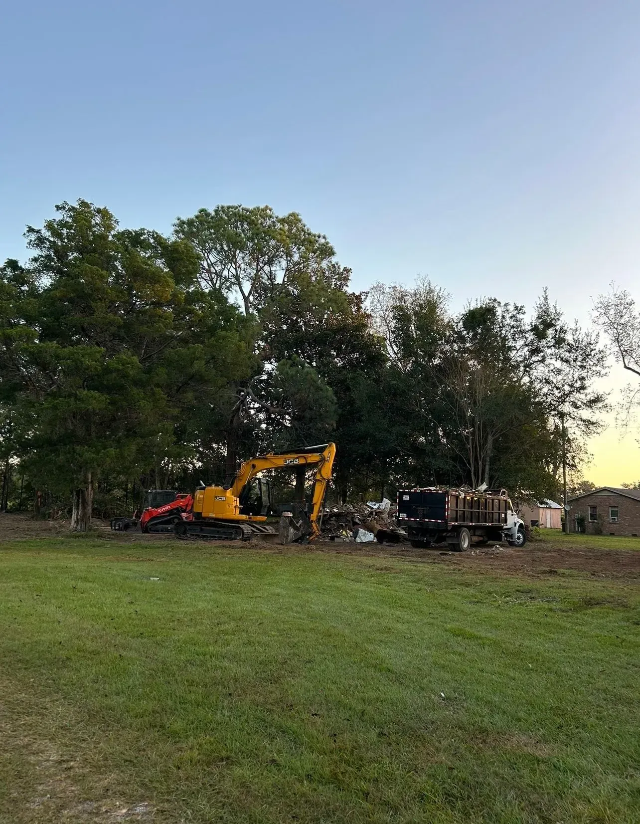 Yellow excavator and dump truck clearing debris on a grassy lot. Trees in the background.