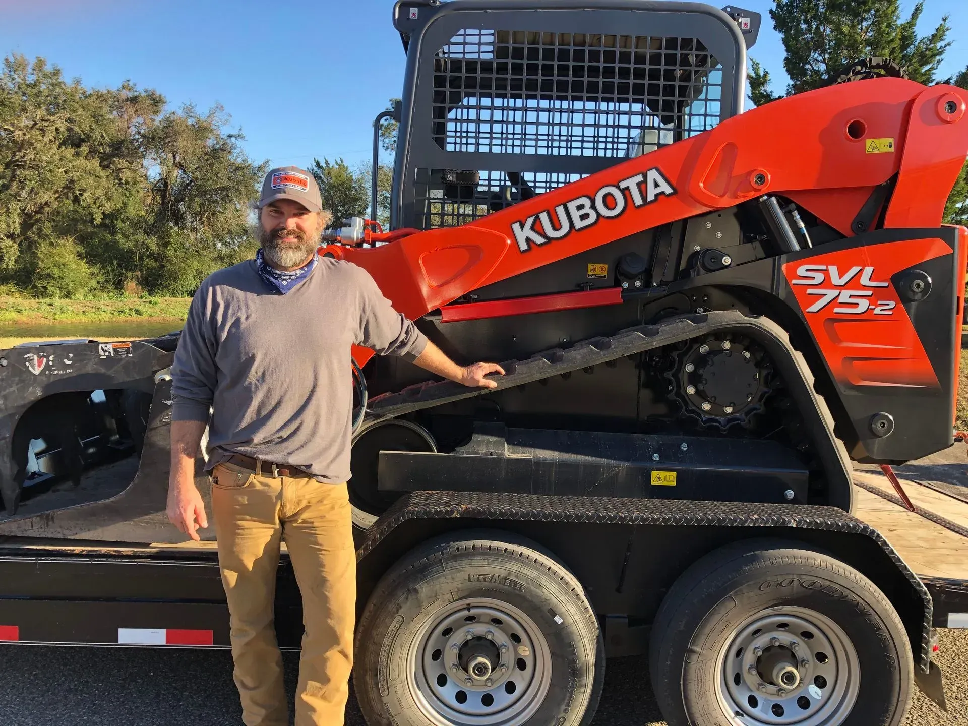 Man stands next to a Kubota SVL75-2 tracked skid steer on a trailer; orange machine and man are outdoors.