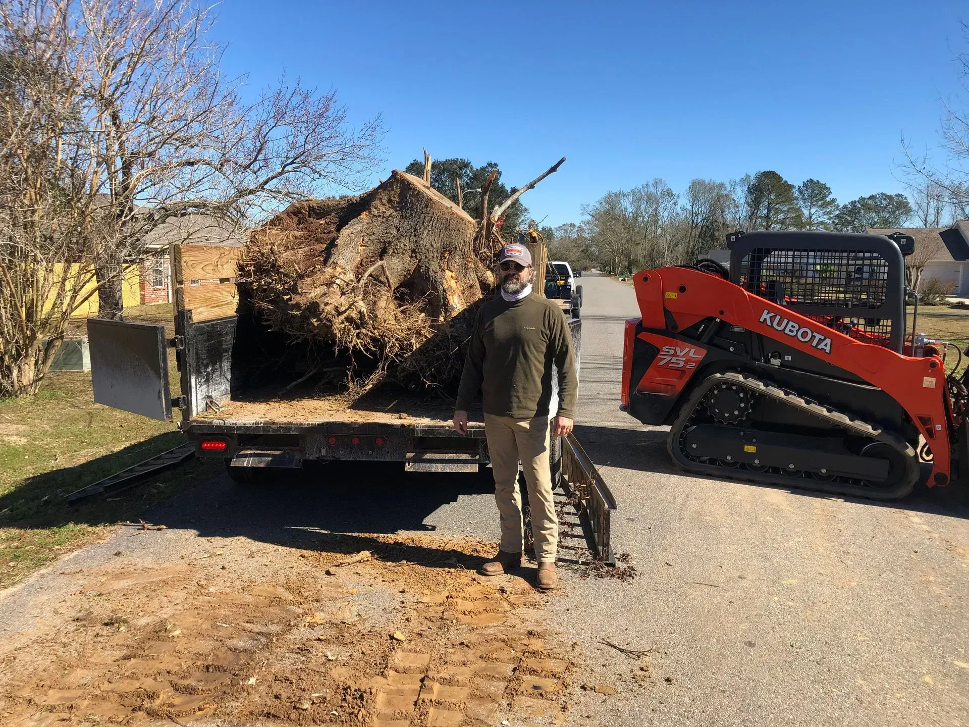 Man standing in front of a truck loaded with tree roots and a Kubota skid steer on a sunny day.