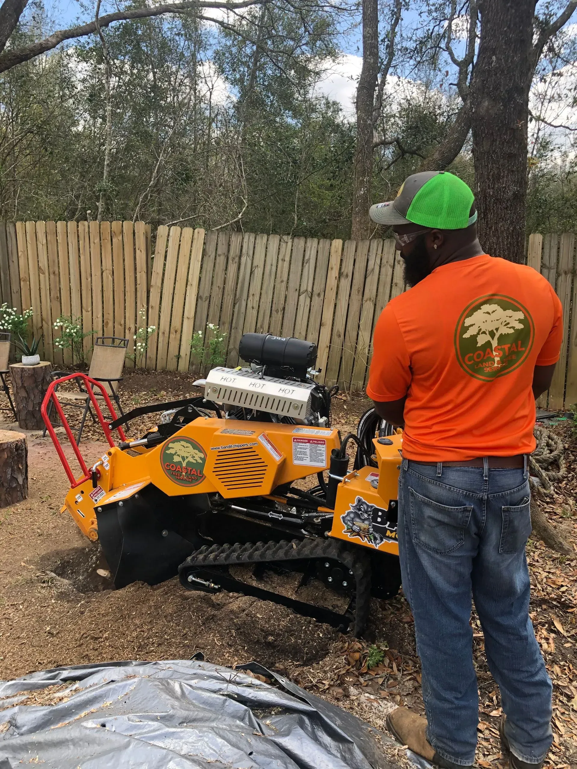 Man operating a yellow stump grinder next to a wooden fence. He wears an orange shirt and a green cap.