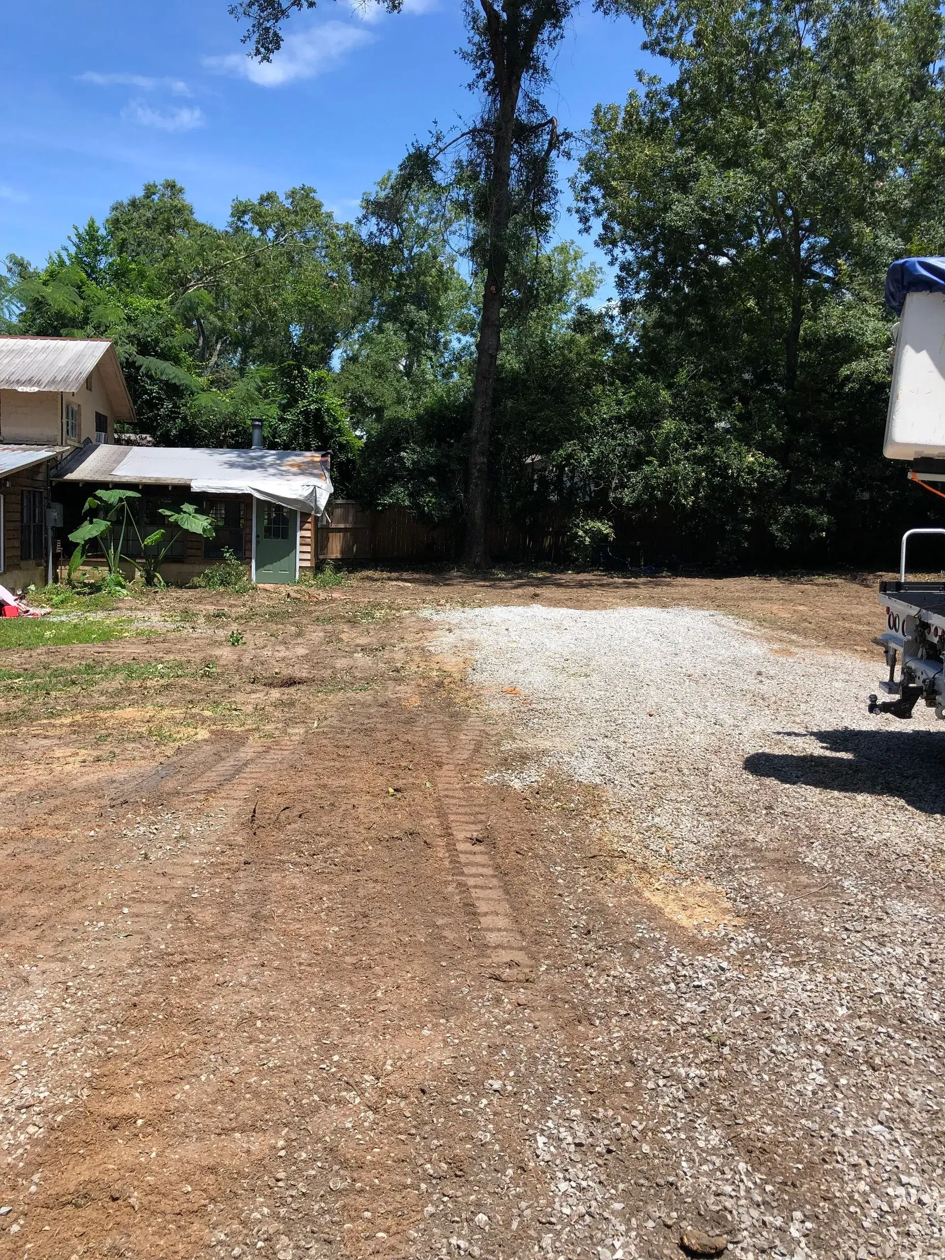 Gravel driveway next to a dirt yard with a small building in the background and trees.