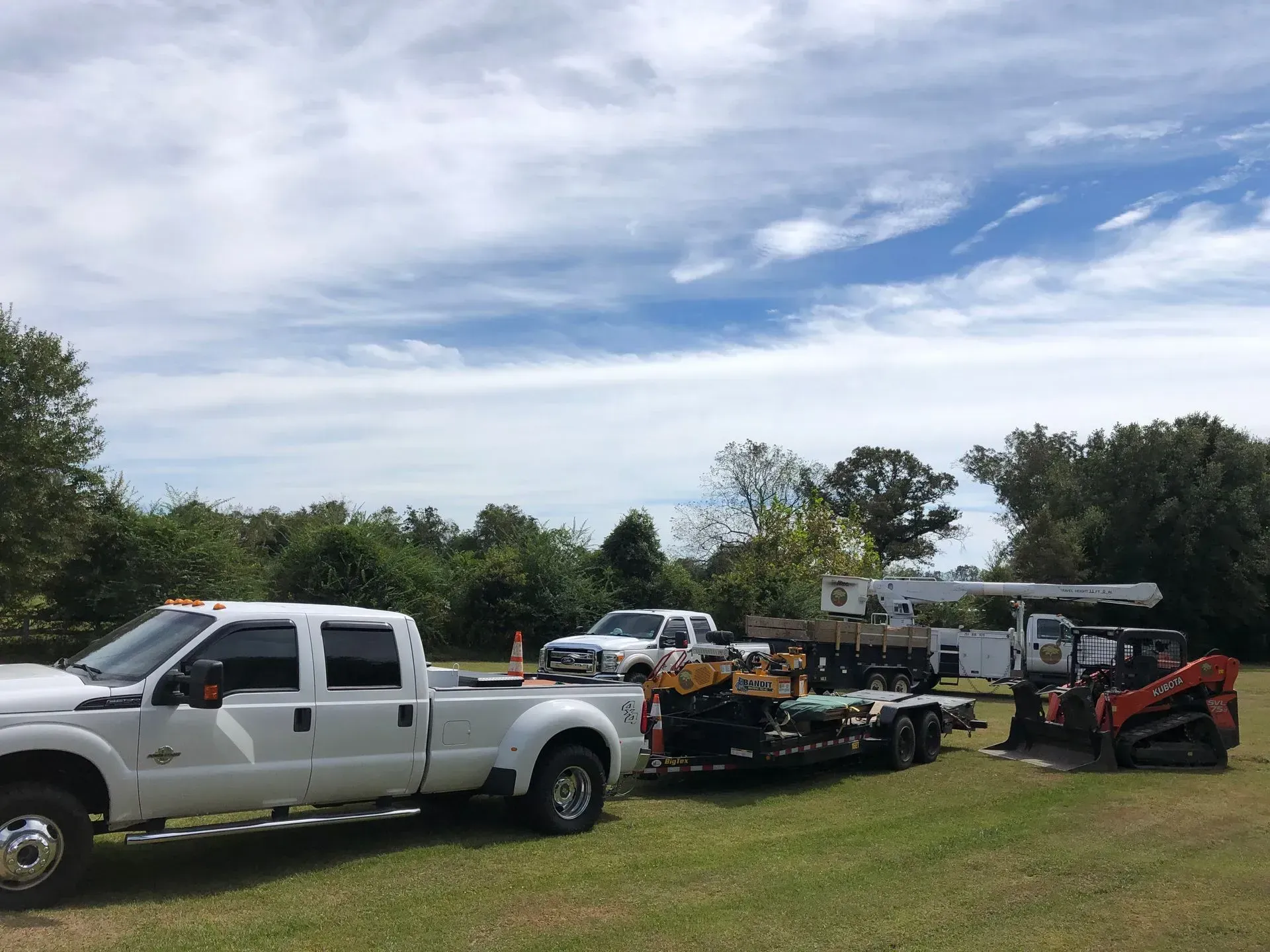 White trucks and equipment trailer on grass under a cloudy sky near trees.