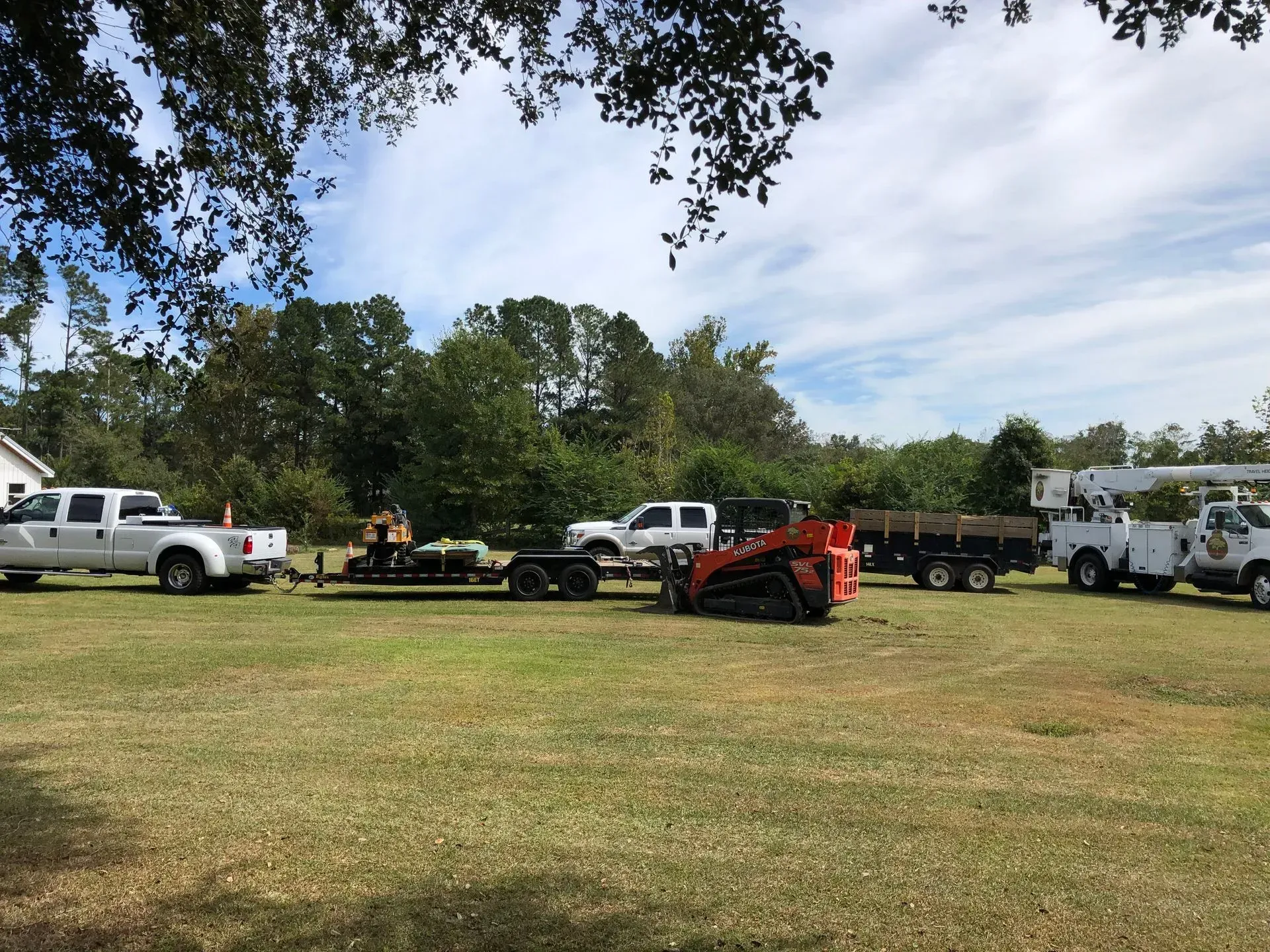 Several work trucks, trailers, and equipment parked on a grassy area near trees under a cloudy sky.