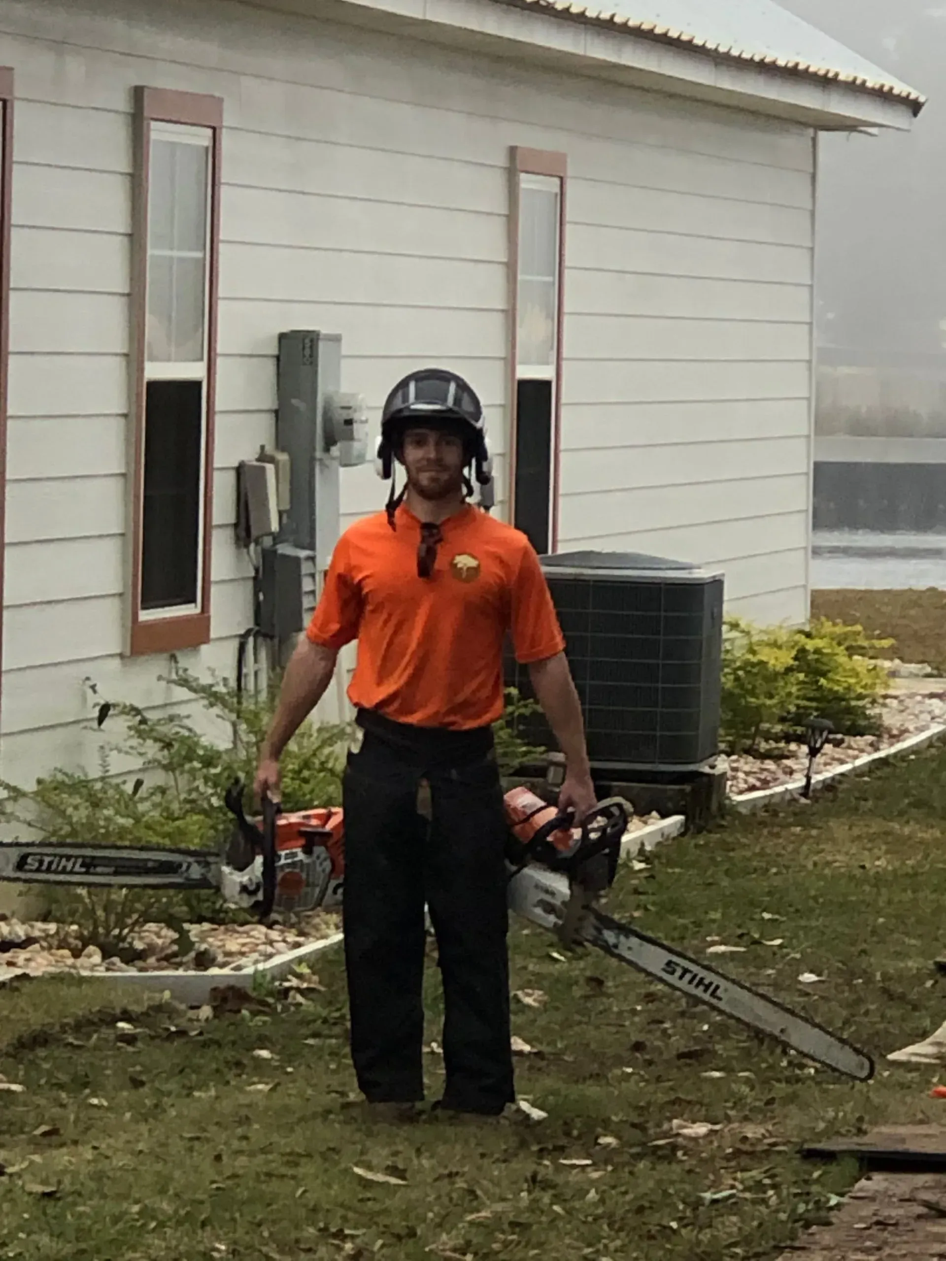 Man wearing helmet holding two chainsaws in front of a white building.
