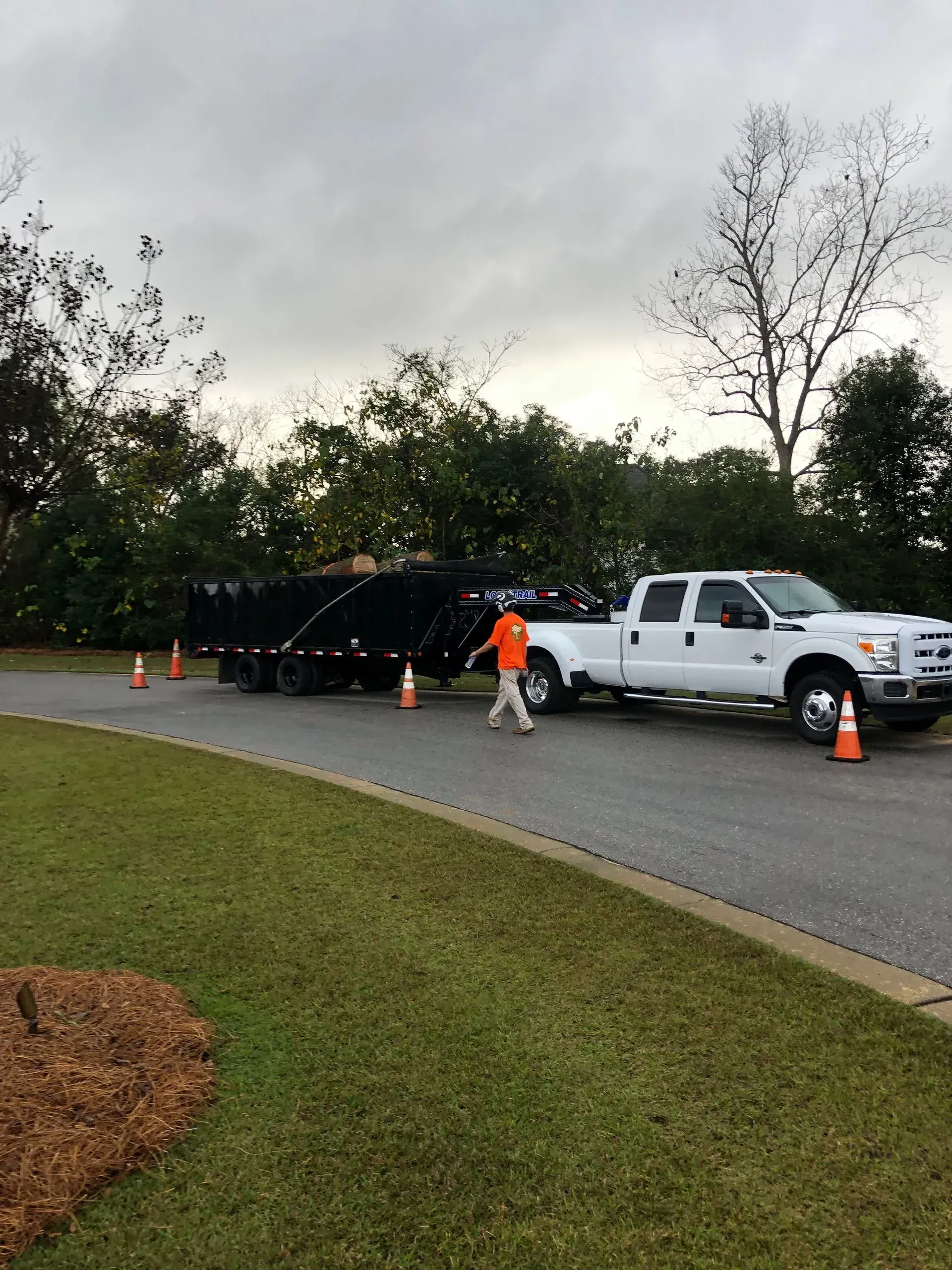 White pickup truck pulling a trailer filled with debris. A person in an orange vest walks on a street with cones.