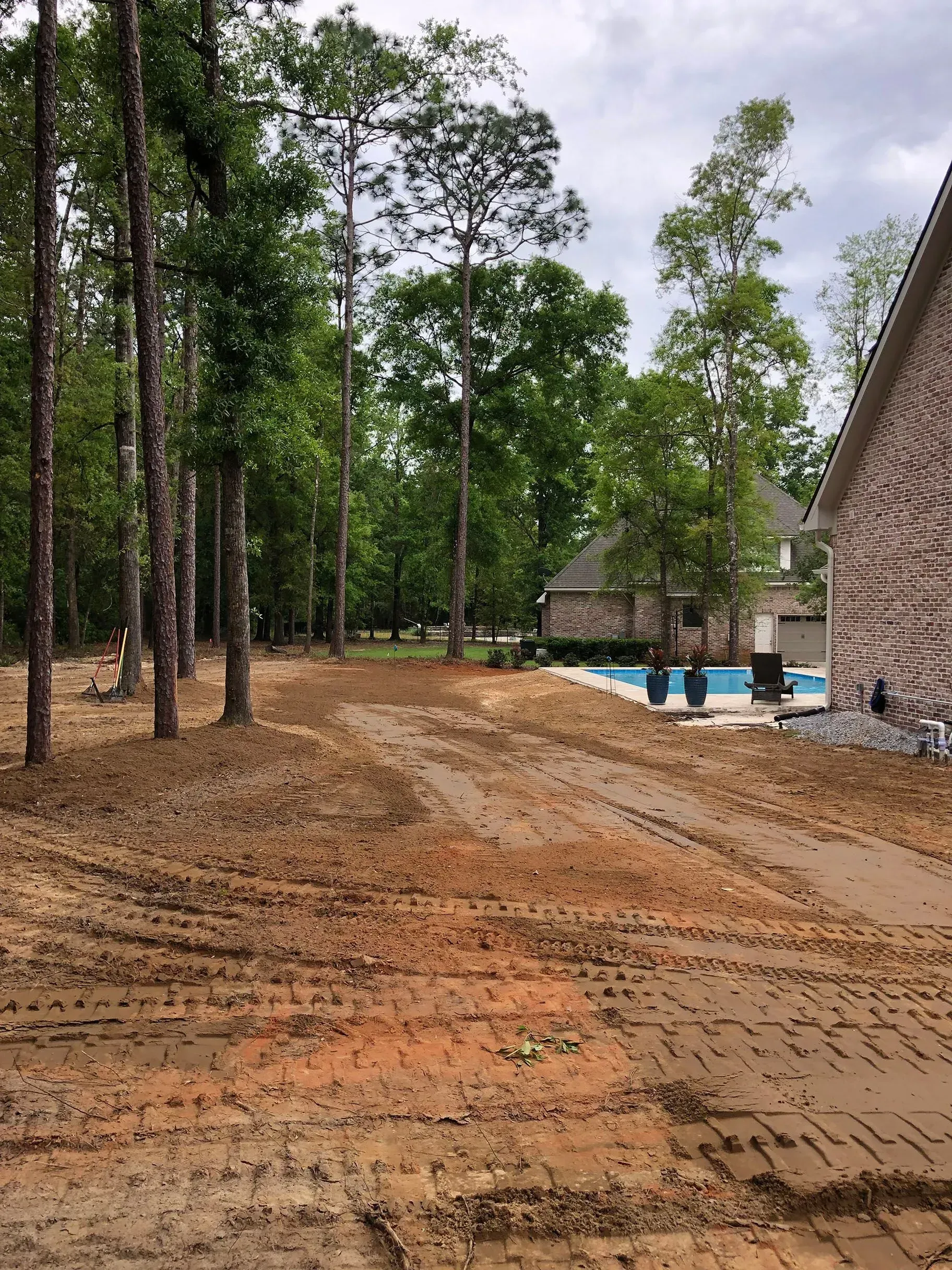 Muddy yard with trees and a house with a pool in the background.