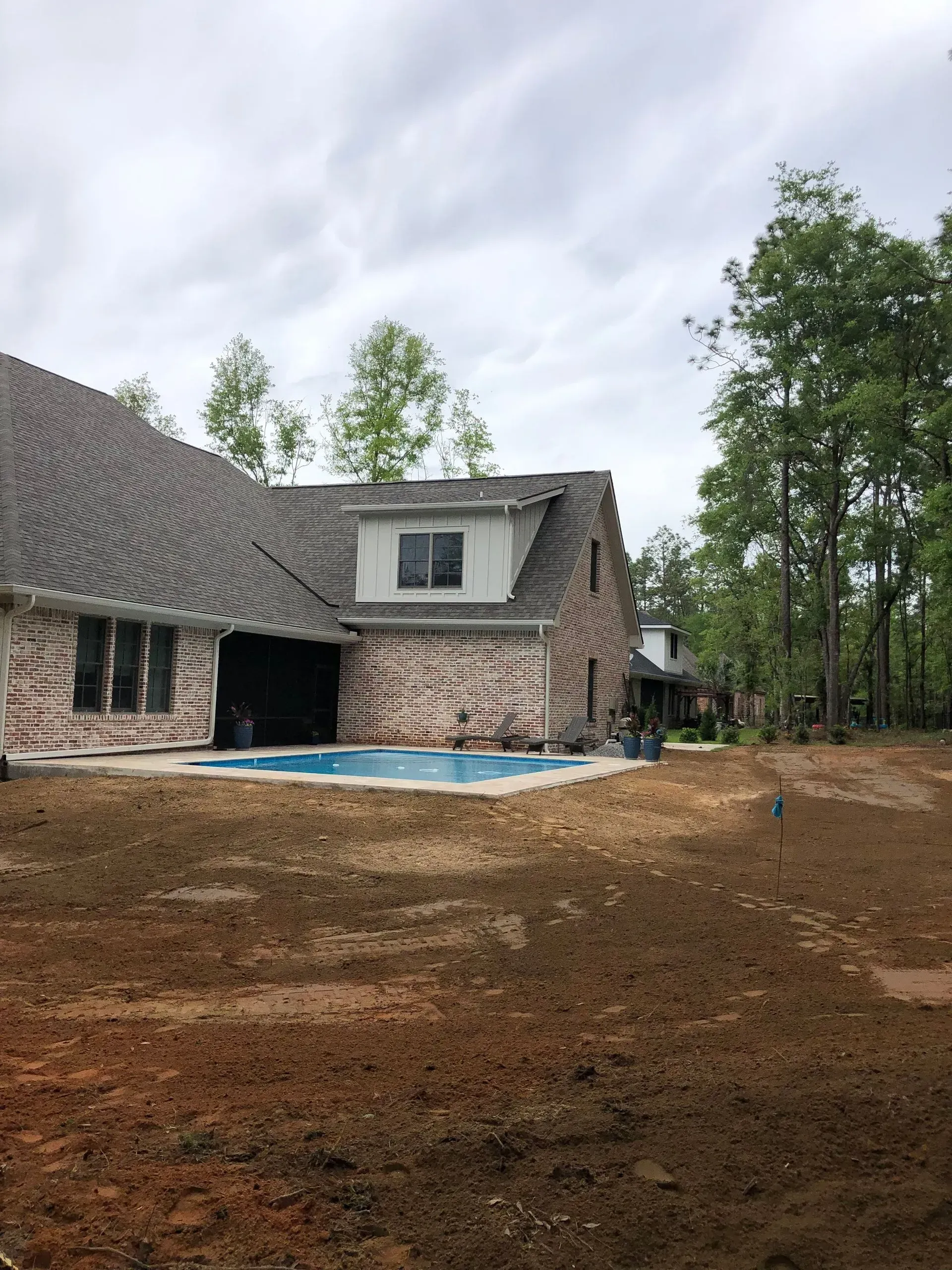 Backyard with house, pool, and trees under a cloudy sky. Brown yard, stone and brick house.