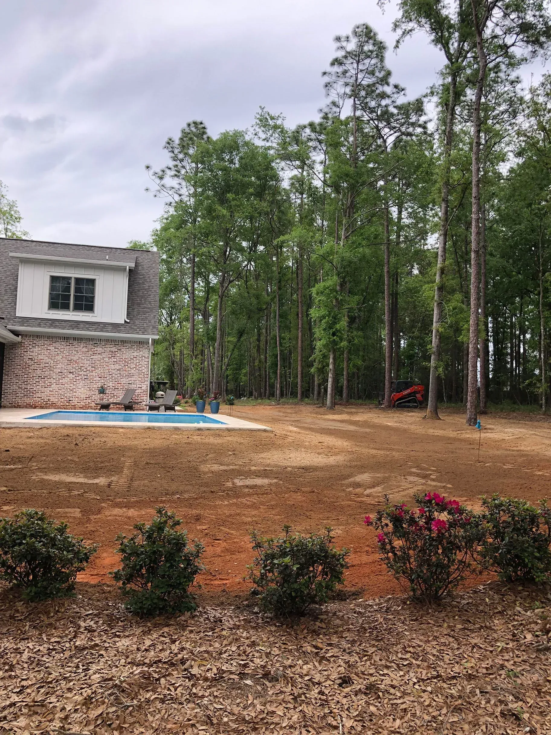 Backyard with a pool, brick house, and a line of trees. Brown yard, cloudy sky.