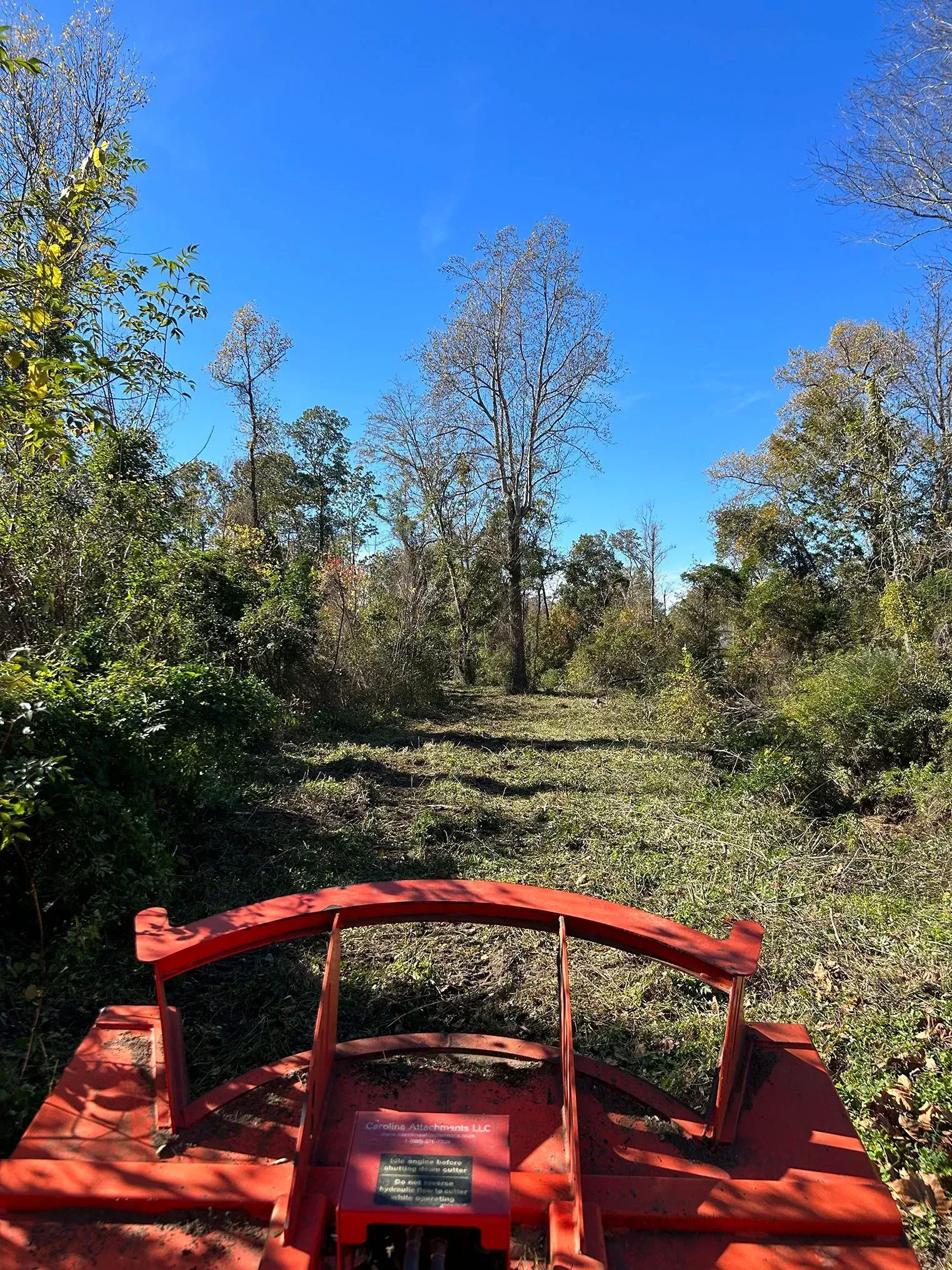 Red brush hog mowing a path through overgrown brush under a blue sky.