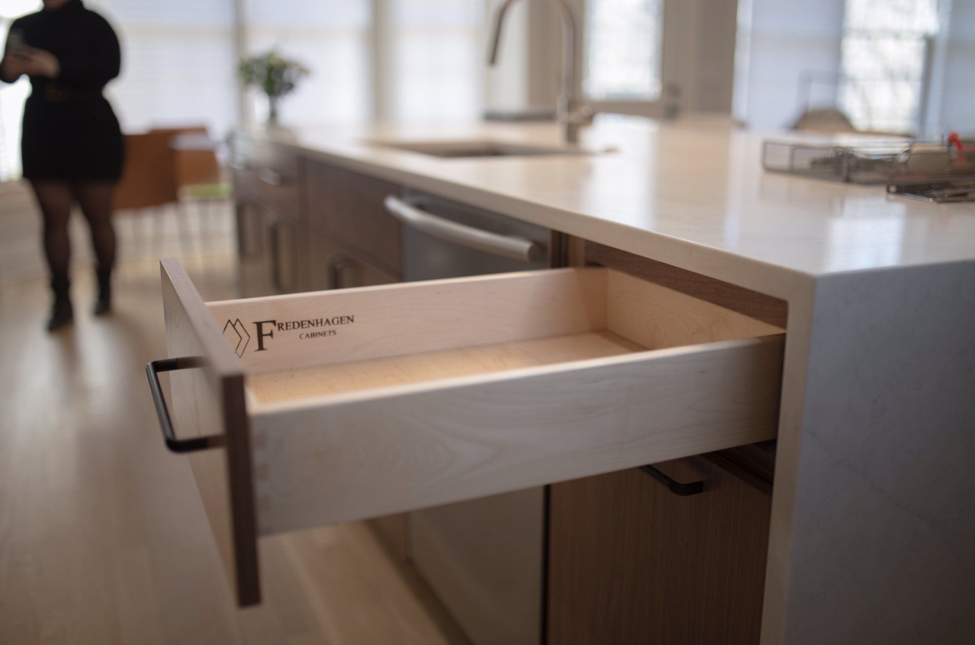 A kitchen with a drawer open and a woman walking in the background.